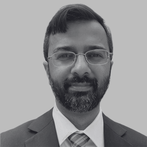 Black and white professional headshot of Farooq Tirmizi, a SovDoc leadership team member, wearing glasses, a suit, and a tie against a neutral background.