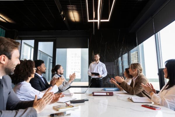 A professional business meeting in a modern conference room, where a presenter in a white shirt speaks to a diverse team applauding and engaging in discussion.